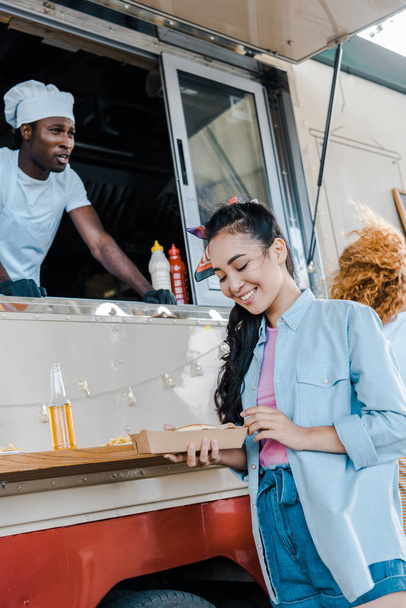selective focus of cheerful asian girl looking at carton plate near chef in food truck  - Photo, Image