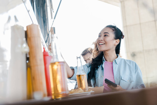 Selektywny fokus szczęśliwy Asian Girl uśmiechając się w pobliżu butelki z piwem i klientami w pobliżu Food Truck  - Zdjęcie, obraz