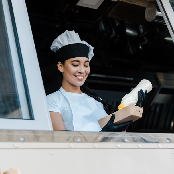 low angle view of happy asian woman holding carton plate and mayonnaise bottle in food truck  - Photo, Image