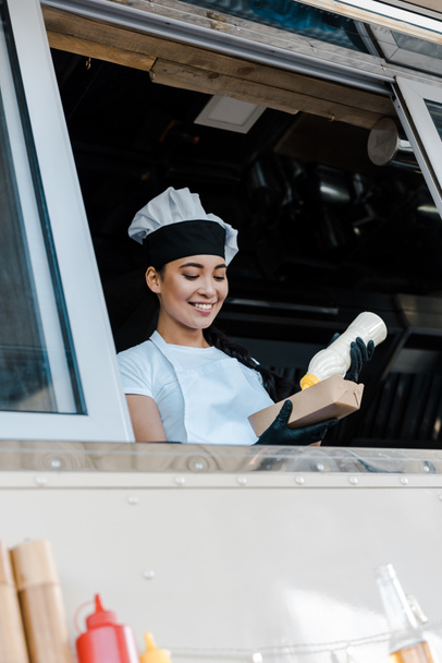 low angle view of cheerful asian woman holding carton plate and mayonnaise bottle in food truck  - Fotoğraf, Görsel
