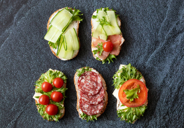 Fresh sandwiches with sausage, cheese, bacon, tomatoes, lettuce, cucumbers on a dark background. Horizontal orientation - Photo, Image