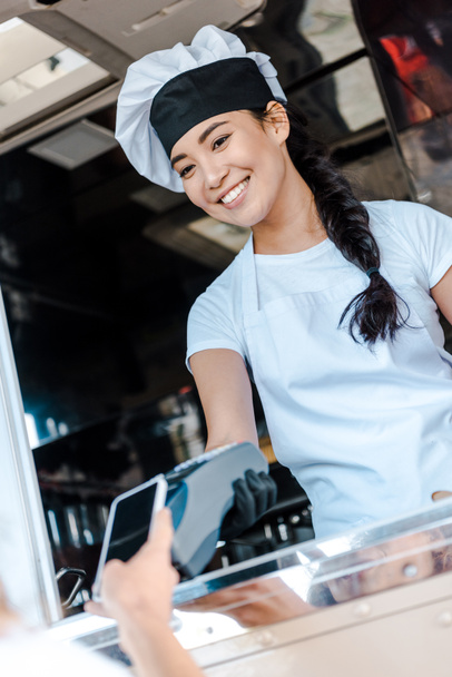 abgeschnittene Ansicht einer Frau, die mit dem Smartphone in der Nähe eines asiatischen Mädchens in einem Food-Truck bezahlt  - Foto, Bild