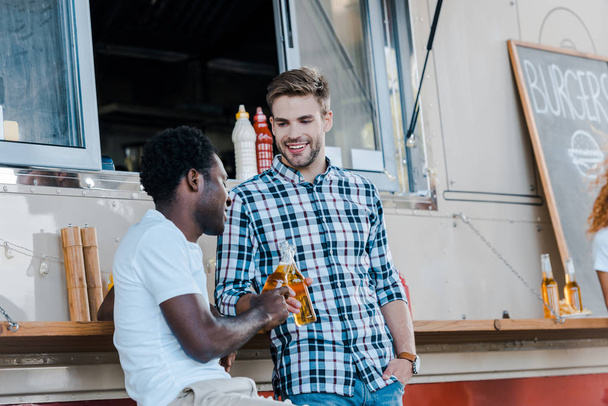 cheerful multicultural men clinking bottles with beer near food truck  - Foto, Bild