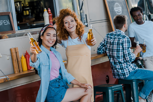 selective focus of happy asian girl holding beer near redhead woman and food truck - Фото, изображение