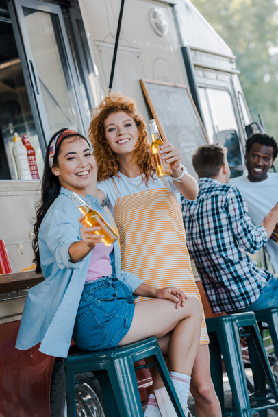 selectieve focus van gelukkige multiculturele meisjes die flessen bier in de buurt van Food truck houden - Foto, afbeelding