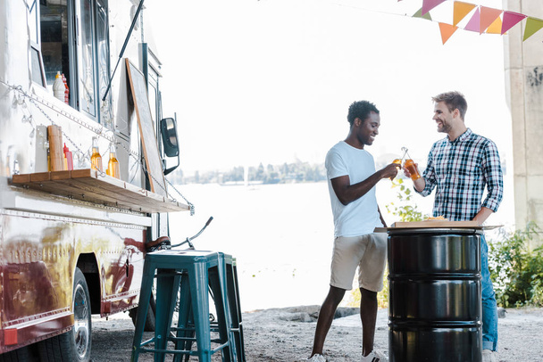 positive multicultural men clinking bottles of beer near food truck outside  - Photo, Image