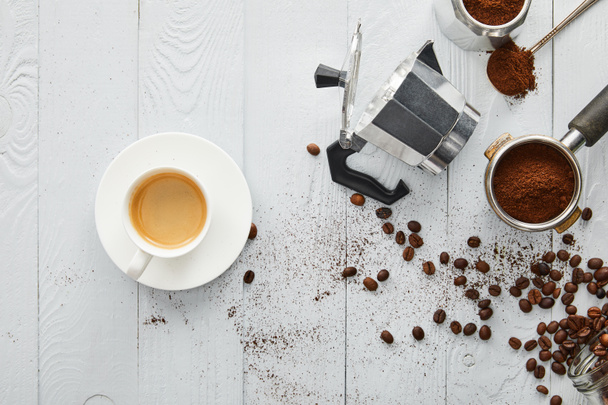 Felülnézet csésze kávét csészealj közelében Gejzer kávéfőző, portafilter és kanalat, fehér fa felületén kávébab - Fotó, kép