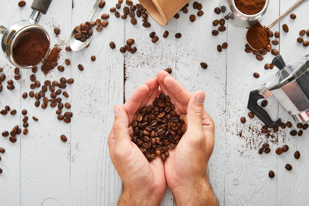 Top view of male hands with coffee beans under white wooden surface with spoons, paper cup, portafilter and geyser coffee maker - Fotografie, Obrázek