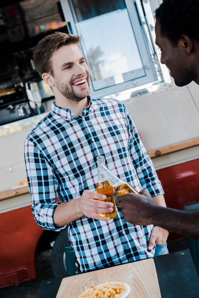 selective focus of cheerful multicultural men toasting bottles of beer near french fries and food truck  - Фото, изображение