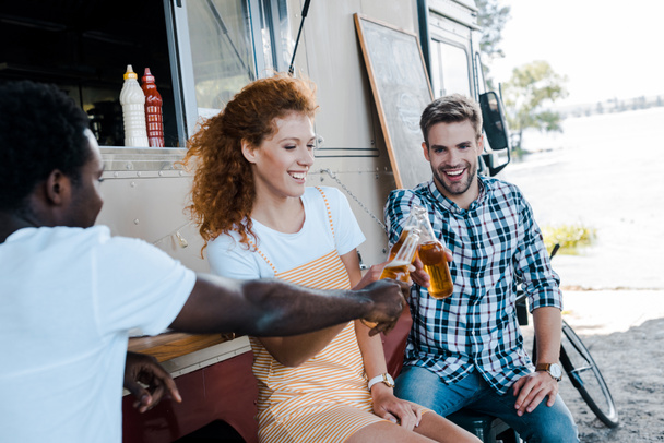 フードトラックの近くに魅力的な赤毛の女性とビールのボトルでクリンキングハンサムな多文化男性の選択的な焦点  - 写真・画像