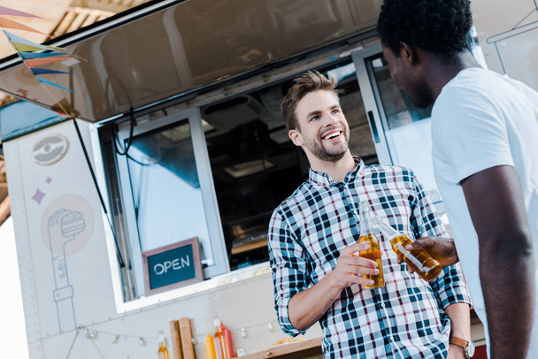 vista de ángulo bajo de amigos multiculturales sonrientes tostando botellas de cerveza cerca de camión de comida
  - Foto, Imagen