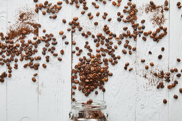 Granos de café dispersos en la superficie de madera blanca con frasco de vidrio
 - Foto, imagen