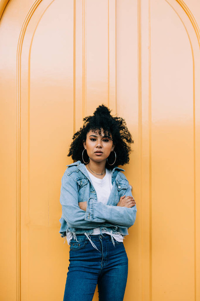 Jonge Afro-Amerikaanse vrouw in jeans en denim jasje leunend op gele deur en kijken naar camera - Foto, afbeelding