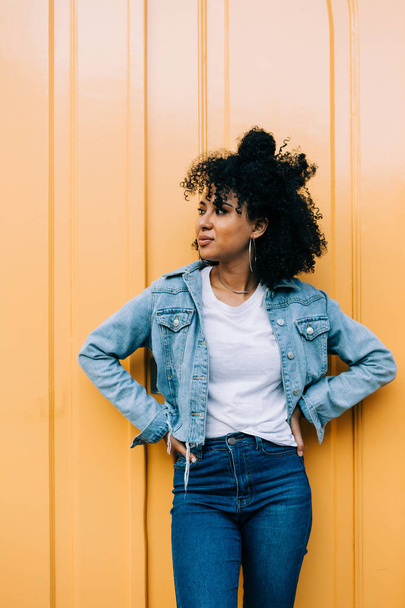Joven mujer afroamericana en jeans y chaqueta de mezclilla apoyada en la puerta amarilla y mirando hacia otro lado
 - Foto, imagen
