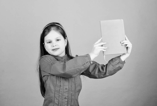 Look at this book. Small pupil took the book from library. Adorable school child with activity book. Cute little girl holding note book with orange cover, copy space - Foto, imagen