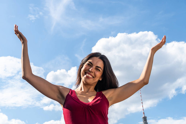 Положительные эмоции: счастливая улыбающаяся брюнетка, позирующая с поднятыми руками на фоне голубого неба
 - Фото, изображение