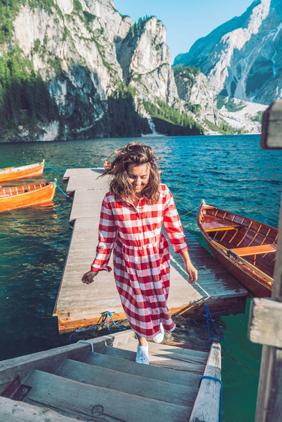 woman walking by mountain lake pier with wooden boats in red dress - Фото, зображення