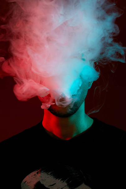 Close-up studio shot of a young bald guy vaping, blowing out a cloud of smoke on red background. - Foto, Bild