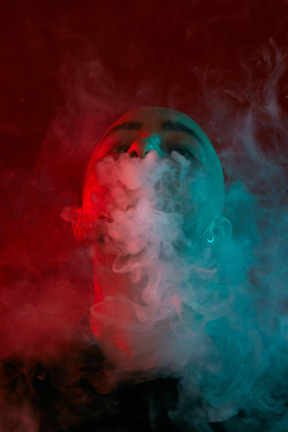 Close-up studio shot of a young bald guy vaping, blowing out a cloud of smoke on red background. - Fotografie, Obrázek