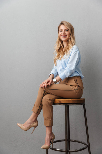 Blonde business woman posing isolated grey wall background sitting on stool. - Foto, Bild