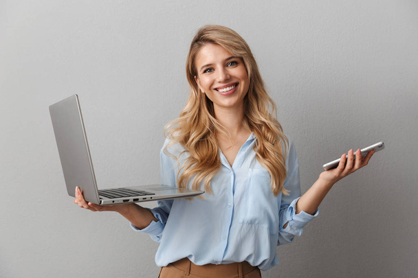 Photo of pleased blonde woman 20s dressed in shirt smiling while holding smartphone and silver laptop - Foto, Bild