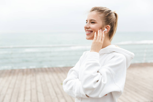 Image of happy nice woman smiling and listening to music with earpods while walking near seaside - Fotografie, Obrázek