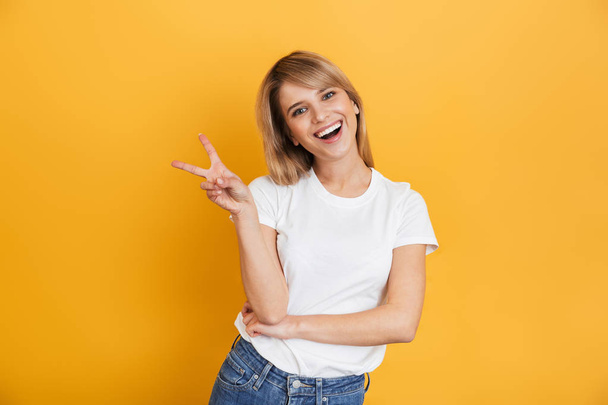 Happy young pretty blonde woman posing isolated over yellow wall background dressed in white casual t-shirt showing peace gesture. - Zdjęcie, obraz
