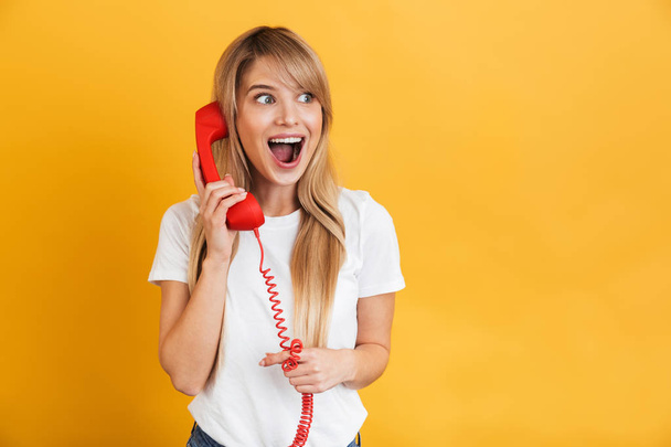 Happy young excited shocked blonde woman posing isolated over yellow wall background talking by retro red telephone. - Foto, immagini