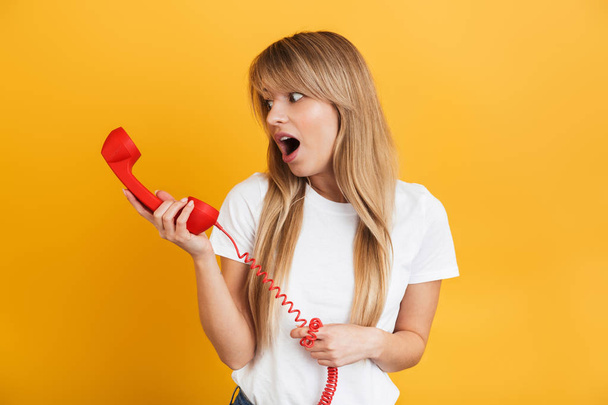 Shocked young blonde woman posing isolated over yellow wall background dressed in white casual t-shirt talking by telephone. - Photo, Image