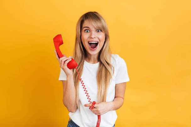 Happy young surprised blonde woman posing isolated over yellow wall background talking by retro red telephone. - Photo, Image