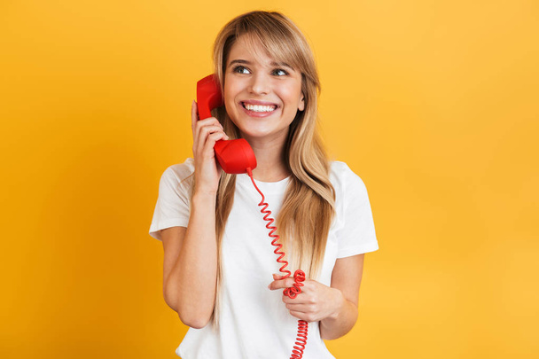 Smiling emotional happy young blonde woman posing isolated over yellow wall background dressed in white casual t-shirt talking by telephone. - Фото, изображение