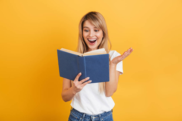 Cheerful emotional young blonde woman posing isolated over yellow wall background dressed in white casual t-shirt holding book reading. - 写真・画像