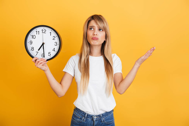 Thinking young blonde woman posing isolated over yellow wall background dressed in white casual t-shirt holding clock. - Foto, immagini