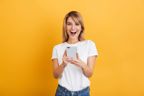 Happy emotional young blonde woman posing isolated over yellow wall background dressed in white casual t-shirt using mobile phone. - Foto, imagen