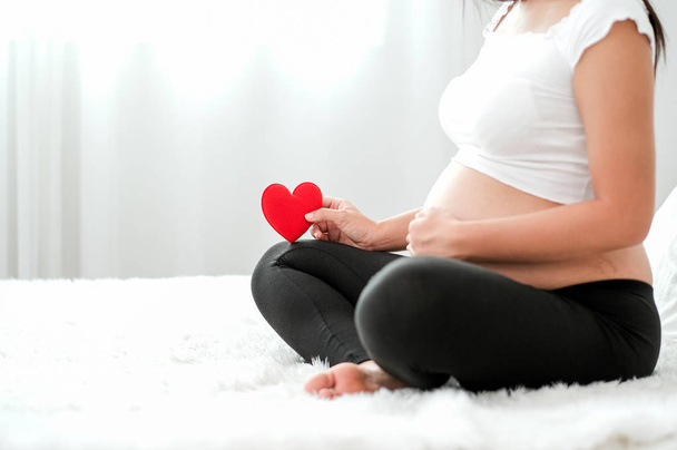 Hamile kadının yan görünümü beyaz yatakta oturup anneden kavram aşk bebek ile karnına veya rahminde kırmızı kalp sembolü tutun. - Fotoğraf, Görsel