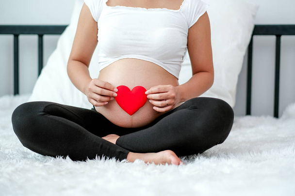 妊娠中の女性の正面図は、白いベッドに座って、母親からの概念愛の赤ちゃんと彼女の腹や子宮に赤いハートのシンボルを保持します. - 写真・画像