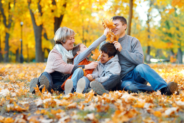 Щаслива сім'я сидить на опале листя, грає і розважається в осінньому міському парку. Діти та батьки разом добре проводять день. Яскраве сонячне світло і жовте листя на деревах, осінній сезон
. - Фото, зображення