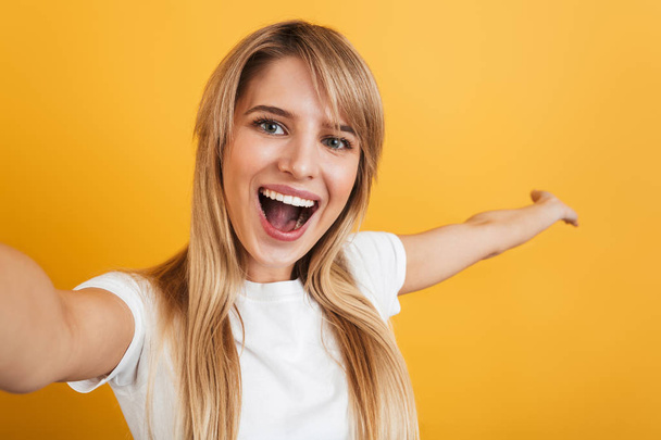 Happy positive pleased young blonde woman posing isolated over yellow wall background dressed in white casual t-shirt take selfie by camera. - Foto, Bild