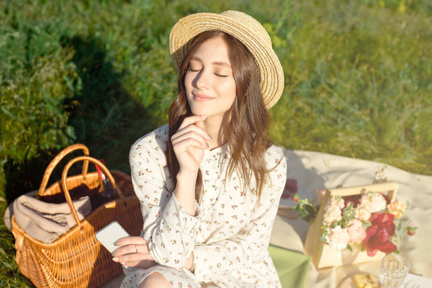 Young beautiful, dark-haired girl in a straw hat at sunset picnic in the field. Autumn. Camping fruits and drinks in a basket and bouquet. - 写真・画像