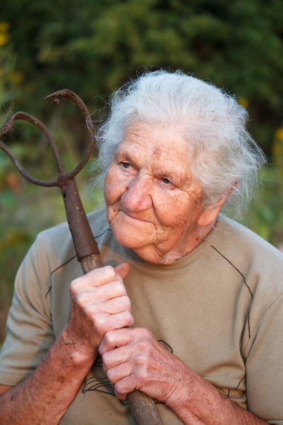 Close-up portrait of an old woman with gray hair holding a rusty pitchfork or chopper in her hands, face in deep wrinkles, selective focus - Photo, Image