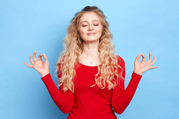 blonde girl dressed in red sweater keeping eyes closed while meditating - Foto, afbeelding