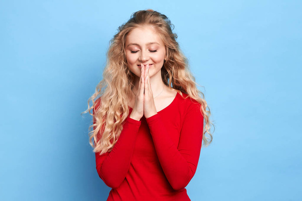 Cute happy cheerful girl with closed eyes keeps hands in praying gesture - Фото, изображение