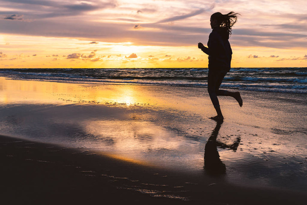 日没時に砂浜のビーチで裸足で走る十代の少女 - 写真・画像
