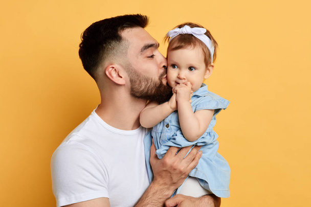 handsome bearded caring father holding and kissinga little girl, isolated on yellow - Photo, Image