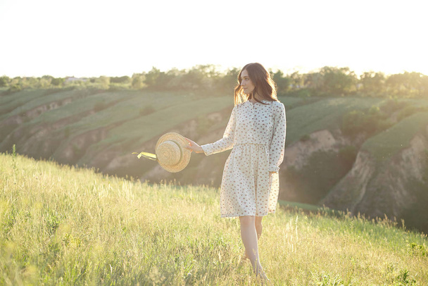 Happy woman with a straw hat in her hands walking on top of the edge of a mountain cliff under the sunset light of the sky, enjoying success, freedom and a bright future. - Foto, imagen