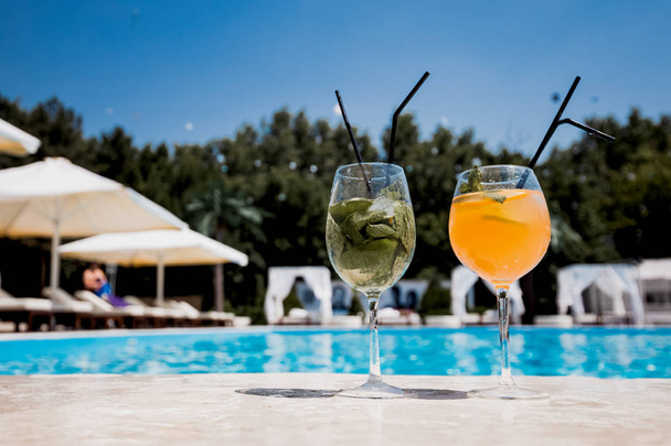 Two glasses with a cocktail stand on the edge of the swimming pool next to lounge zone on the open air on a sunny summer day - Foto, Imagem