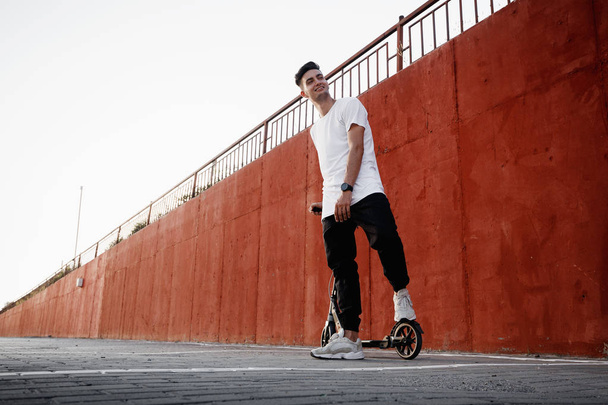 Young guy dressed in jeans and t-shirt stands with a scooter against a painted concrete wall on the summer day in the city. - Photo, image