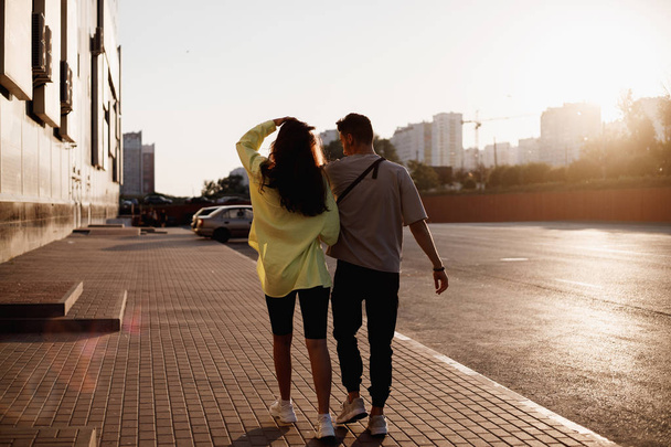 Young stylish guy and girl are walking together on the square for parking next to the building on the sunset - 写真・画像