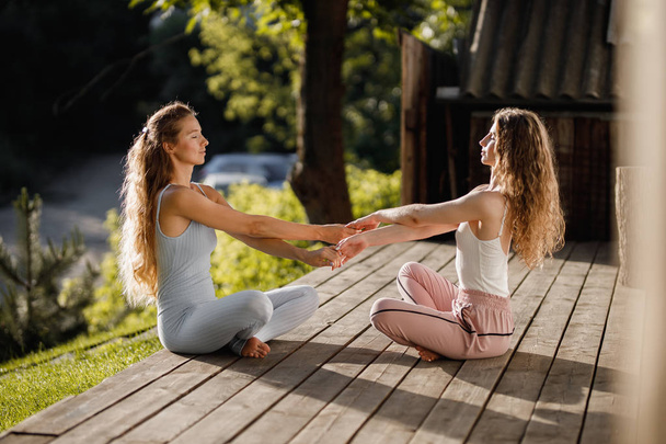 Two young women are sitting and holding hands in the lotus position on a wooden podium in the garden near the house in the sunny morning - Фото, изображение