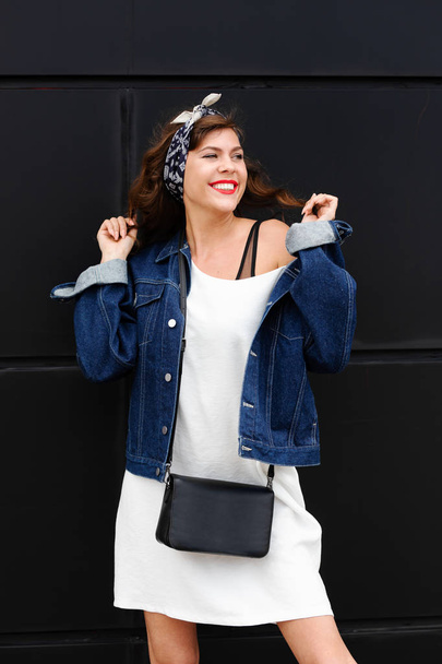 Stylish young girl in trendy casual clothes in a denim jacket and headband poses in the street on a summer day - Φωτογραφία, εικόνα
